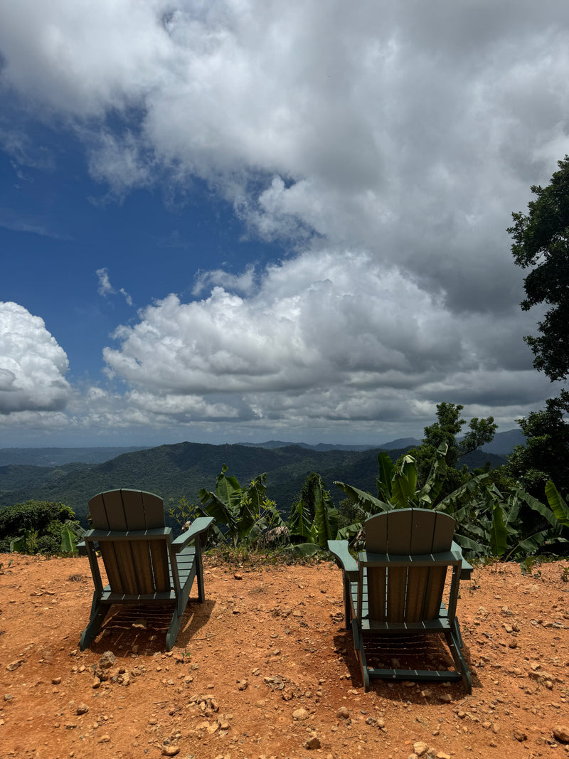 Two wooden chairs on a dirt patch overlooking a scenic landscape with mountains and clouds from finca vegas arriba, puerto rico.