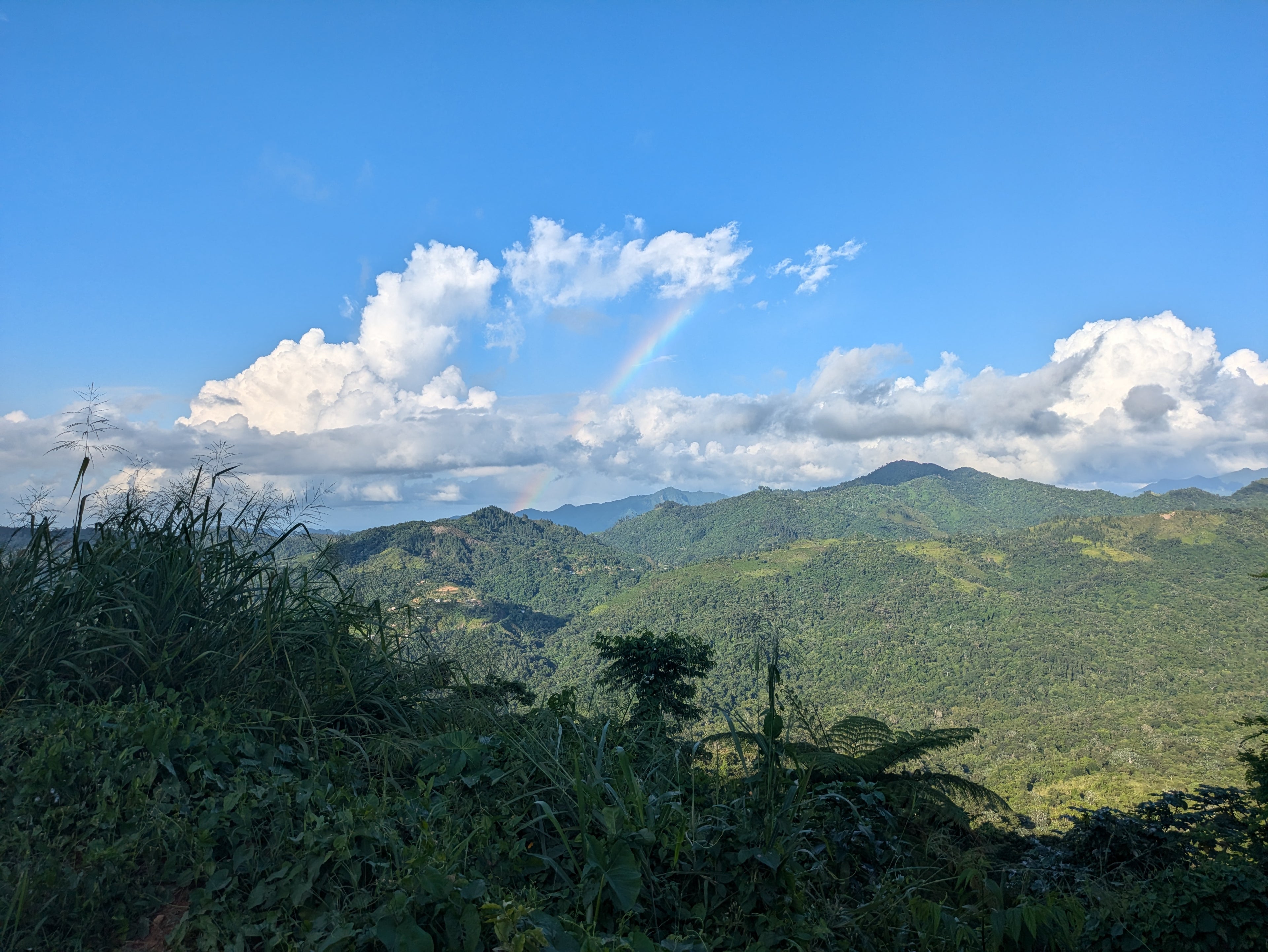 Scenic view of green mountains of Adjuntas, Puerto Rico under a blue sky with clouds and a rainbow. Finca Vegas Arriba sky view.